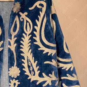 Handmade deep teal blue velvet suzani jacket with large-scale cream floral and paisley embroidery, open front with striped lining and flower button closure artisan boho outerwear for women, displayed on a hanger against a warm beige wall.