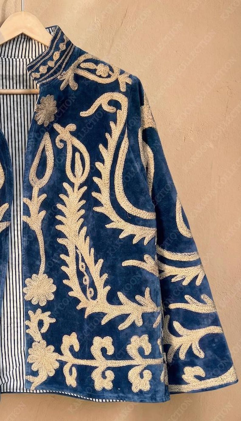 Handmade deep teal blue velvet suzani jacket with large-scale cream floral and paisley embroidery, open front with striped lining and flower button closure artisan boho outerwear for women, displayed on a hanger against a warm beige wall.