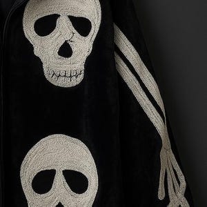 May include: Black jacket with embroidered white skull and bone designs. The jacket features two large skulls on the front and a skeletal arm on the sleeve. The garment is hanging on a wooden hanger against a dark background.