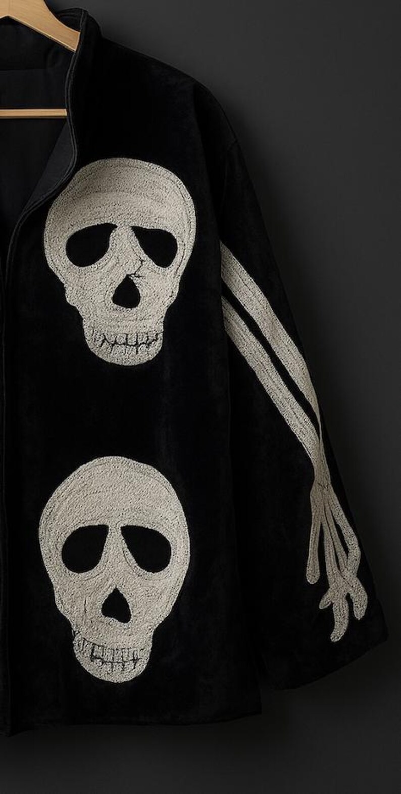 May include: Black jacket with embroidered white skull and bone designs. The jacket features two large skulls on the front and a skeletal arm on the sleeve. The garment is hanging on a wooden hanger against a dark background.