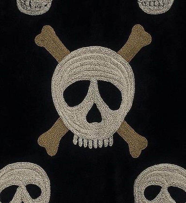 May include: Black fabric with an embroidered skull and crossbones design. The skull is white with a detailed texture, and the crossbones are tan. Several skull and crossbones motifs are visible across the fabric.