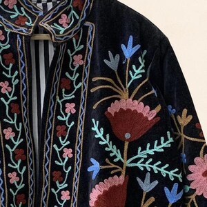 May include: A close-up of a dark blue velvet jacket with colorful floral embroidery. The jacket features pink, red, and blue flowers with green leaves. The collar and edges are also embroidered with a floral design. The inside lining has a black and white striped pattern.