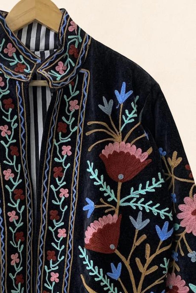 May include: A close-up of a dark blue velvet jacket with colorful floral embroidery. The jacket features pink, red, and blue flowers with green leaves. The collar and edges are also embroidered with a floral design. The inside lining has a black and white striped pattern.