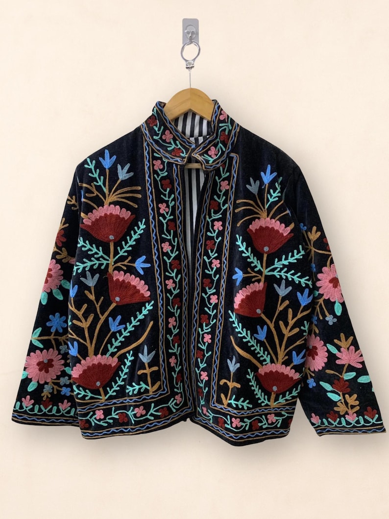 May include: A black velvet jacket with colorful floral embroidery. The jacket features a high collar and is adorned with red, pink, blue, and gold floral patterns. The interior lining has a striped pattern. The jacket is hanging on a wooden hanger.