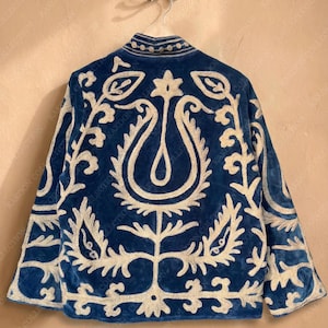 Handmade deep teal blue velvet suzani jacket with large-scale cream floral and paisley embroidery, open front with striped lining and flower button closure artisan boho outerwear for women, displayed on a hanger against a warm beige wall.