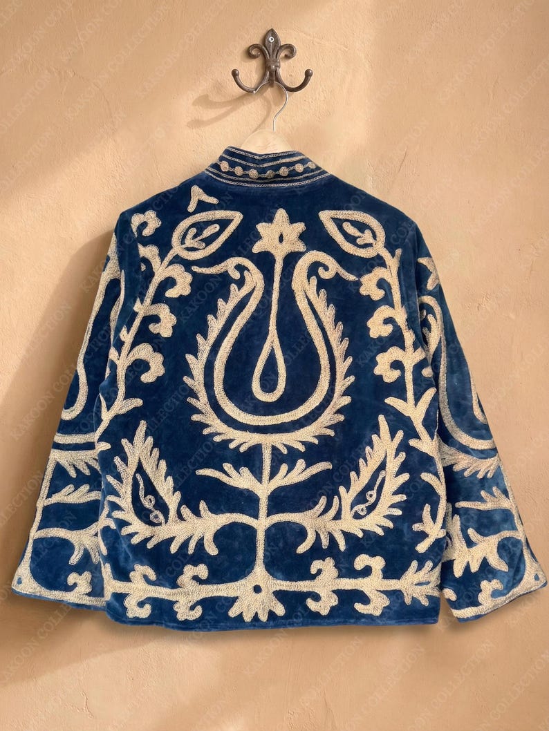 Handmade deep teal blue velvet suzani jacket with large-scale cream floral and paisley embroidery, open front with striped lining and flower button closure artisan boho outerwear for women, displayed on a hanger against a warm beige wall.