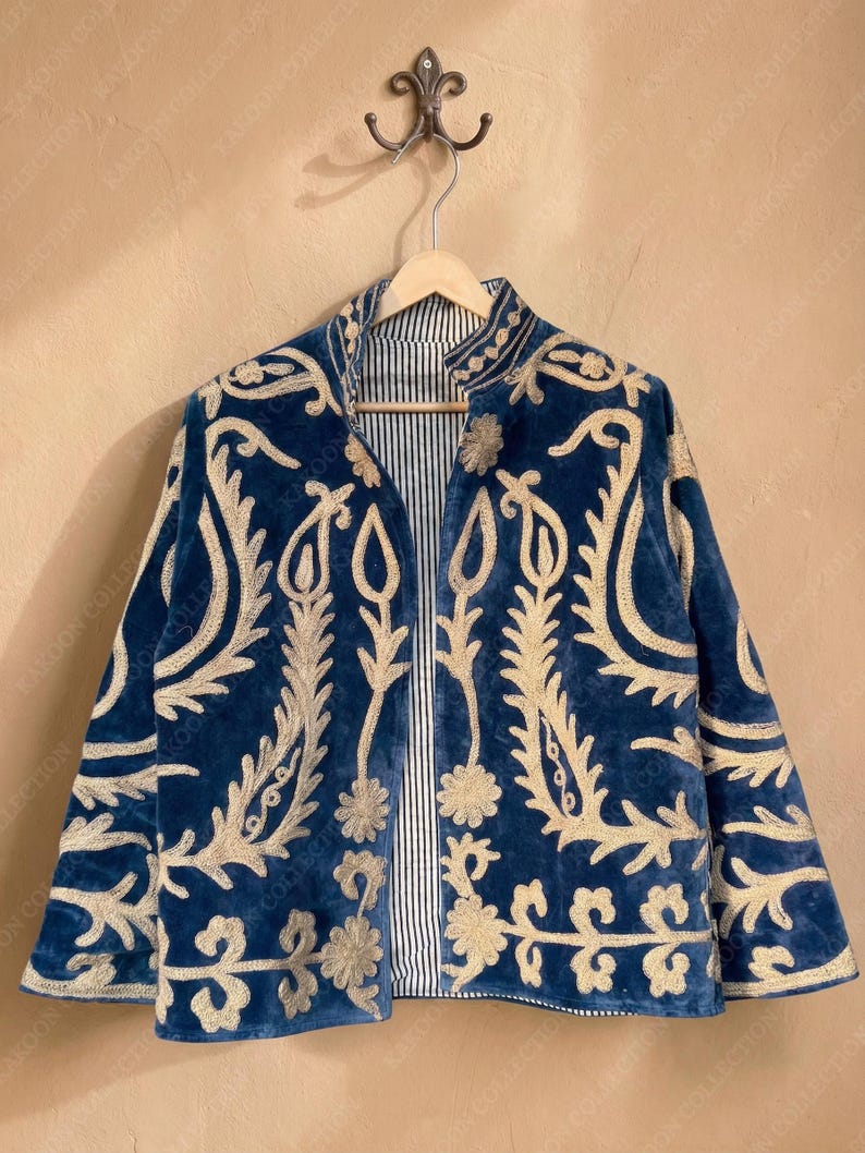 Handmade deep teal blue velvet suzani jacket with large-scale cream floral and paisley embroidery, open front with striped lining and flower button closure artisan boho outerwear for women, displayed on a hanger against a warm beige wall.