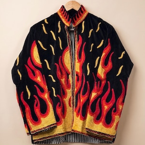 May include: A black jacket featuring an embroidered flame pattern in shades of red, orange, and yellow. The garment has a high collar, long sleeves, and a black and white vertical striped lining visible at the front opening and the bottom hem.