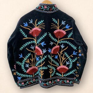 May include: Black velvet jacket with colorful floral embroidery. The jacket features a high collar and intricate embroidery in shades of red, pink, blue, and gold. The floral design is symmetrical and covers the back of the jacket.