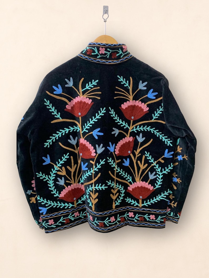 May include: Black velvet jacket with colorful floral embroidery. The jacket features a high collar and intricate embroidery in shades of red, pink, blue, and gold. The floral design is symmetrical and covers the back of the jacket.
