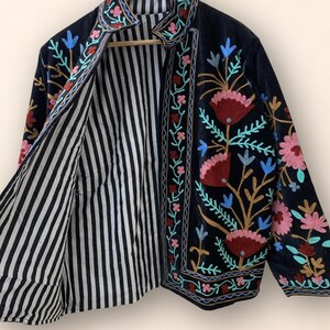 May include: A black jacket with floral embroidery in pink, red, blue, and green. The jacket's interior features a black and white striped pattern. The jacket is hanging on a wooden hanger.