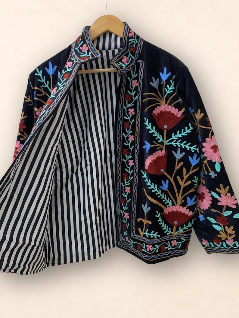 May include: A black jacket with floral embroidery in pink, red, blue, and green. The jacket's interior features a black and white striped pattern. The jacket is hanging on a wooden hanger.
