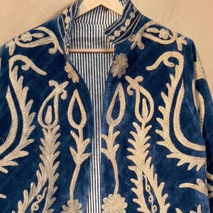 Handmade deep teal blue velvet suzani jacket with large-scale cream floral and paisley embroidery, open front with striped lining and flower button closure artisan boho outerwear for women, displayed on a hanger against a warm beige wall.