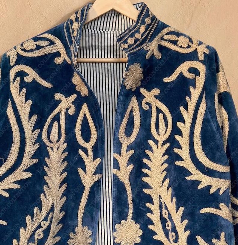 Handmade deep teal blue velvet suzani jacket with large-scale cream floral and paisley embroidery, open front with striped lining and flower button closure artisan boho outerwear for women, displayed on a hanger against a warm beige wall.