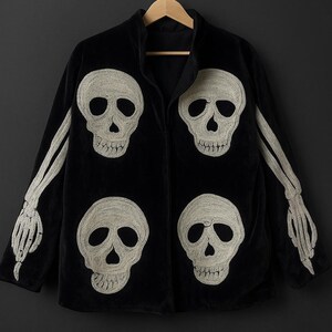 May include: Black jacket with embroidered white skulls and skeleton arms. The jacket features four skull designs on the front and skeleton arms extending from the sleeves. The jacket is hanging on a wooden hanger against a dark background.