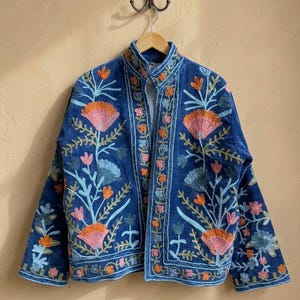 May include: A blue embroidered jacket with a mandarin collar. The jacket features a floral design in shades of orange, pink, and blue. The embroidery is detailed and covers the front and sleeves of the jacket. The jacket is hanging on a wooden hanger.