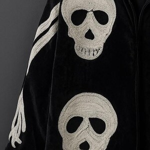 May include: Black velvet jacket with embroidered white skull designs. The jacket features two large skulls on the front and a bone design on the sleeve. The jacket has a full zipper and a hood.
