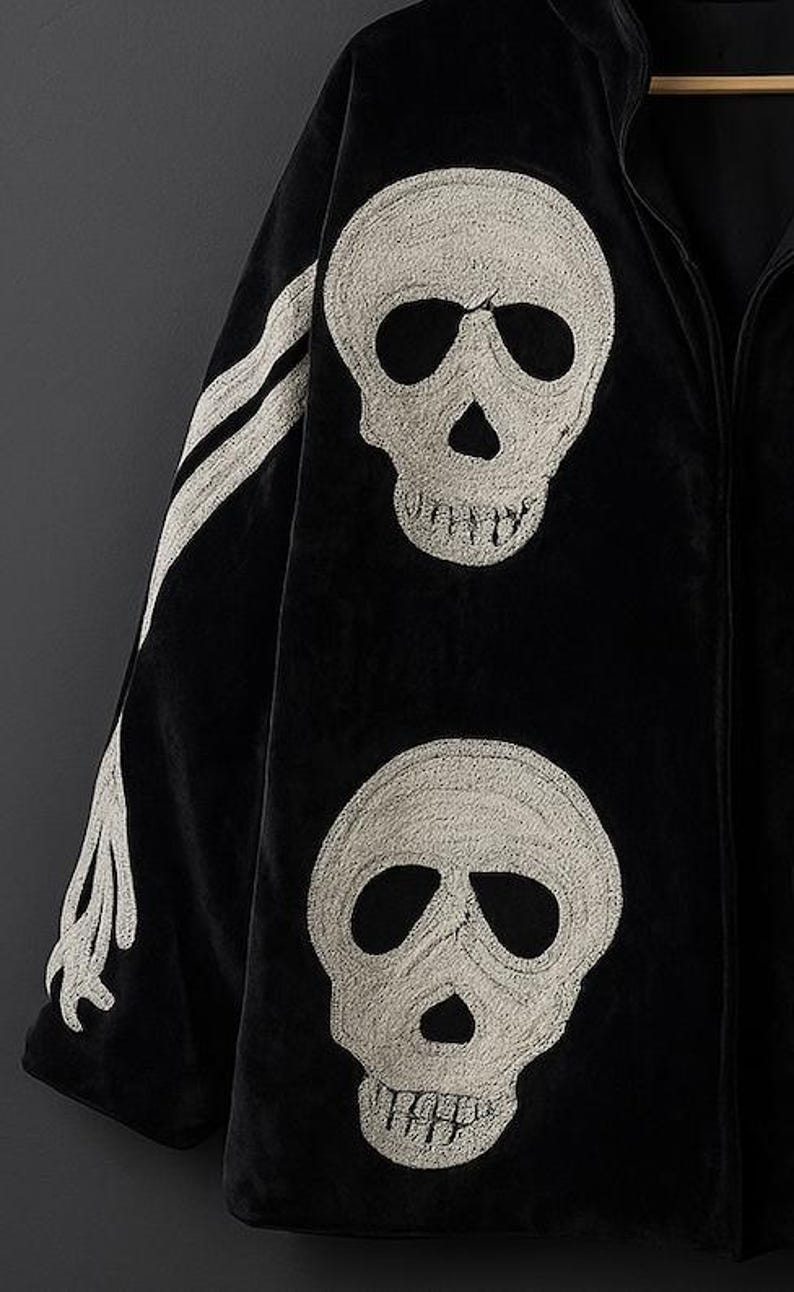May include: Black velvet jacket with embroidered white skull designs. The jacket features two large skulls on the front and a bone design on the sleeve. The jacket has a full zipper and a hood.