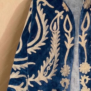 Handmade deep teal blue velvet suzani jacket with large-scale cream floral and paisley embroidery, open front with striped lining and flower button closure artisan boho outerwear for women, displayed on a hanger against a warm beige wall.