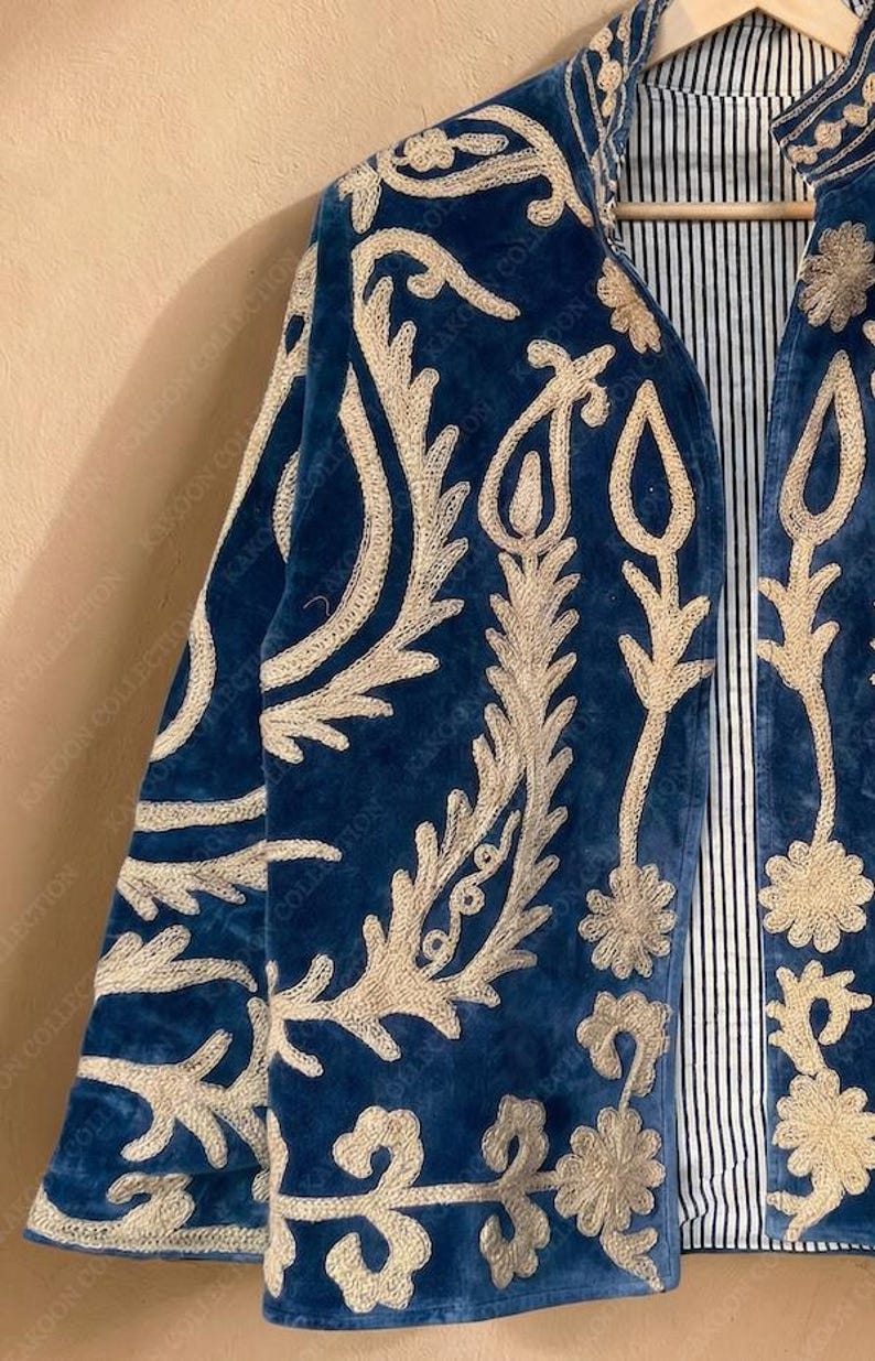 Handmade deep teal blue velvet suzani jacket with large-scale cream floral and paisley embroidery, open front with striped lining and flower button closure artisan boho outerwear for women, displayed on a hanger against a warm beige wall.