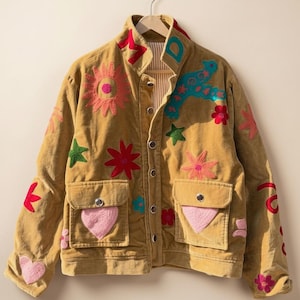 May include: A tan corduroy jacket featuring a button-up front, two front pockets with flaps, and colorful embroidered patches including flowers, stars, hearts, and letters. The jacket has a high collar and long sleeves with additional floral embroidery.