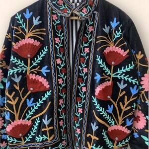 May include: Black velvet jacket with colorful floral embroidery. The jacket features a collar and a striped lining. The embroidery includes red, pink, blue, and gold thread, creating a vibrant and detailed design.