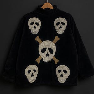May include: Black turtleneck sweater with embroidered skull and crossbones design. The sweater features five embroidered skulls in a cream color, with a larger skull and crossbones in the center. The sweater is hanging on a wooden hanger.