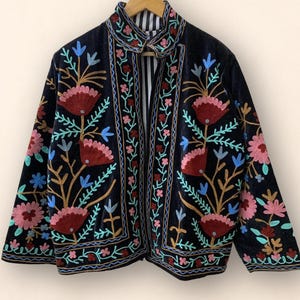 May include: Black velvet jacket with colorful floral embroidery. The jacket features a high collar and a striped lining. The embroidery includes red, pink, blue, and gold thread, creating a vibrant and detailed design.