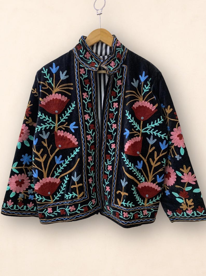 May include: Black velvet jacket with colorful floral embroidery. The jacket features a high collar and a striped lining. The embroidery includes red, pink, blue, and gold thread, creating a vibrant and detailed design.