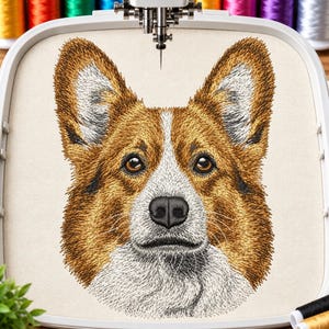 May include: A detailed machine embroidery design of a Corgi dog face centered in a white plastic embroidery hoop. The dog features golden-brown and white fur, dark brown eyes, and a black nose, stitched onto a light beige fabric background.