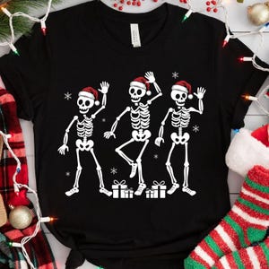 Può includere: T-shirt nero con un design a tema natalizio. Presenta tre figure di scheletri bianchi con cappelli da Babbo Natale che salutano. Il design include fiocchi di neve e scatole regalo. Una maglietta grafica divertente e festosa.