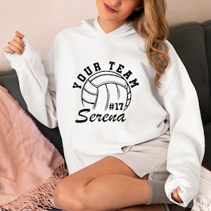 Puede incluir: Sudadera con capucha blanca con un diseño de voleibol negro. El diseño incluye las palabras "YOUR TEAM", una ilustración de voleibol, el número "17" y el nombre "Serena". La sudadera tiene una capucha con cordón y un bolsillo delantero.