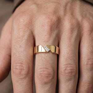 May include: A polished gold band ring featuring a diagonal gap in the center. A single, clear, rectangular baguette-cut gemstone is set within this angled opening, creating a modern, asymmetrical design across the smooth, metallic surface.