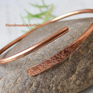 Könnte beinhalten: Ein kupferfarbenes Armreif-Armband mit gehämmerten Texturen an einem Ende. Das offene Design ermöglicht eine einfache Anpassung. Das Armband wird auf einem grauen Stein präsentiert, mit dem Text "HandmadeArtsOnline" sichtbar.