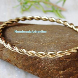 以下が含まれることがあります： 金色のねじれたバングルブレスレット。ブレスレットは、ロープのようなデザインを作るためにねじれた金属の連続したストランドでできています。「HandmadeArtsonline」というテキストが表示されます。