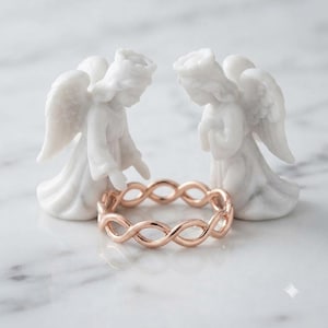 May include: A rose gold ring featuring an intricate infinity or twisted rope design, positioned between two small, white, matte-finished angel figurines with detailed wings and robes, standing upright in a symmetrical arrangement.