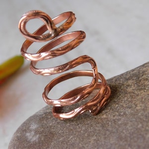 Può includere: Un anello a spirale color rame, con un'asola aperta in alto e fasce strutturate. L'anello è esposto su una pietra grigia, con un fiore arancione e giallo sfocato sullo sfondo. Il design dell'anello presenta motivi intricati.