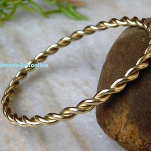 May include: A gold-coloured, twisted metal bangle bracelet. The bracelet has a continuous, rope-like design. The background includes a stone and a marble surface. The text "HandmadeArtsOnline" is visible.