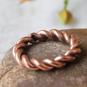 May include: A copper-coloured ring with a twisted rope design. The ring is made of a shiny, reddish-brown metal and sits on a smooth, brown stone. The ring's design features intertwined strands.