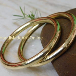 Op de afbeelding: Twee goudkleurige armbanden liggen op een bruine steen tegen een witte achtergrond. De armbanden zijn cirkelvormig en hebben een glad, gepolijst oppervlak. De tekst "HandMadeArtsOnline" is zichtbaar.