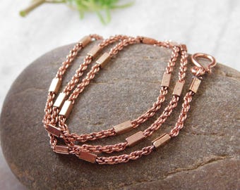 May include: A rose gold necklace with three strands of delicate chain. Each strand features rectangular bar accents. The necklace is displayed on a gray stone, with a clasp visible at the end.