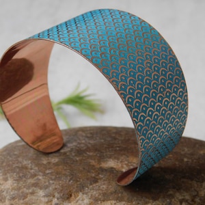 Pode incluir: Pulseira de punho de cobre com um padrão de escamas turquesa e cobre. A pulseira aberta é exibida em uma pedra. A pulseira tem aproximadamente 5 cm de largura.