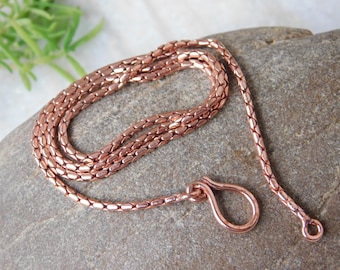 May include: A copper-colored chain necklace with a unique, textured design. The necklace is made of small, interconnected links, and it is displayed on a gray stone surface. The chain has a loop closure.