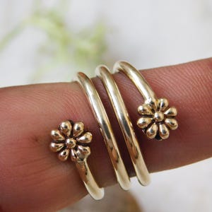 May include: A gold-colored ring wraps around a finger, featuring a spiral design. The ring has three bands and is adorned with two daisy-shaped floral accents. The ring is a piece of jewelry.