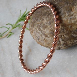 May include: A copper-colored, twisted metal bracelet. The bracelet is circular and has a braided appearance. The bracelet is resting on a light-colored surface next to a smooth, brown rock and a sprig of green leaves.