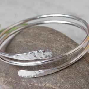 Könnte beinhalten: Ein silberfarbenes Armreif-Armband mit gehämmertem Finish. Das Armband hat ein Spiraldesign mit zwei Bändern und einem offenen Ende. Die gehämmerten Textur verleiht dem Schmuck ein einzigartiges Detail. Das Armband liegt auf einem grauen Stein.