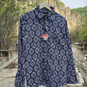 Op de afbeelding: Een overhemd met lange mouwen en knopen, versierd met een marineblauw en wit paisley patroon. Het overhemd hangt aan een houten hanger. Een label met een cartoonfiguur is aan het overhemd bevestigd. Het overhemd is gemaakt van lichte stof.