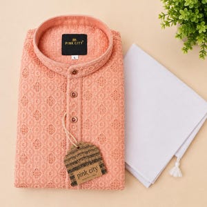 K&ouml;nnte beinhalten: Ein pfirsichfarbener Langarm-Kurta mit einem komplizierten gestickten geometrischen Muster, einem Stehkragen und einer Knopfleiste. Ein schwarzes Etikett zeigt PINK CITY mit der Gr&ouml;&szlig;e L. Ein schlichtes, wei&szlig;es, gefaltetes Stoffst&uuml;ck liegt bei.