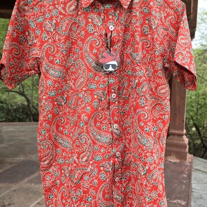 May include: Red short-sleeved button-down shirt with a paisley pattern in teal and white. The shirt is hanging on a wooden hanger. The tag reads "PINK CITY".