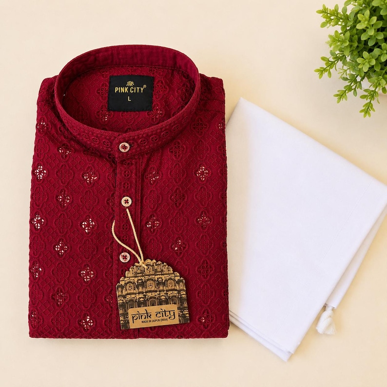 K&ouml;nnte beinhalten: Ein dunkelroter Langarm-Kurta mit Stehkragen, Knopfleiste und einem aufwendigen Lochstickereimuster. Ein Etikett mit der Aufschrift PINK CITY ist am Hemd befestigt, das mit einem gefalteten wei&szlig;en Kleidungsst&uuml;ck kombiniert ist.